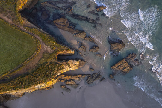 Aerial view of As Catedrais beach featuring dramatic rock formations, sandy shoreline, and turquoise waves crashing against the cliffs in Spain.