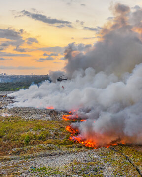 Aerial view of a helicopter fighting a large fire at the Vertedero Duquesa landfill with thick smoke rising into the sunset sky in Pantoja, Santo Domingo, Dominican Republic.