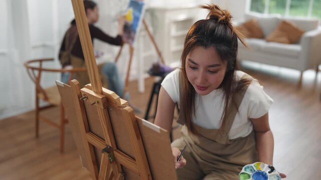 Creative young Asian women friends painting on canvas together in bright home studio. Relaxed female artists enjoying peaceful indoor leisure time, expressing imagination and passion for art.