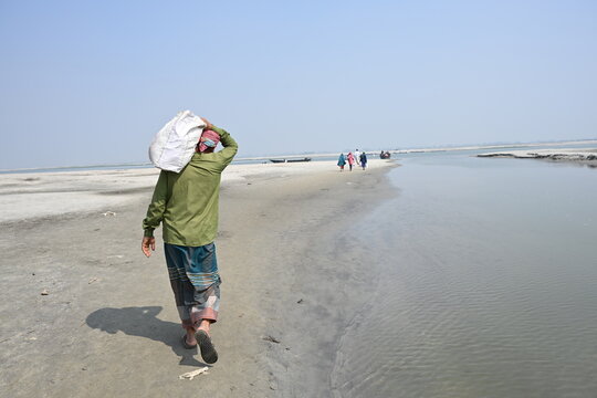 Gaibandha District, Rangpur Division, Bangladesh - 15 July 2026: View of a man carrying a sack on his head while walking along the sandy banks of Char Krishnamani with other people in the distance.