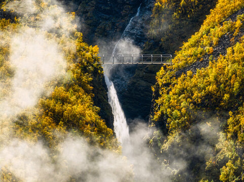 Aerial view of Gorsa bridge spanning a deep canyon with a powerful waterfall, surrounded by vibrant autumn foliage and rising mist in Birtavarre, Troms, Norway.