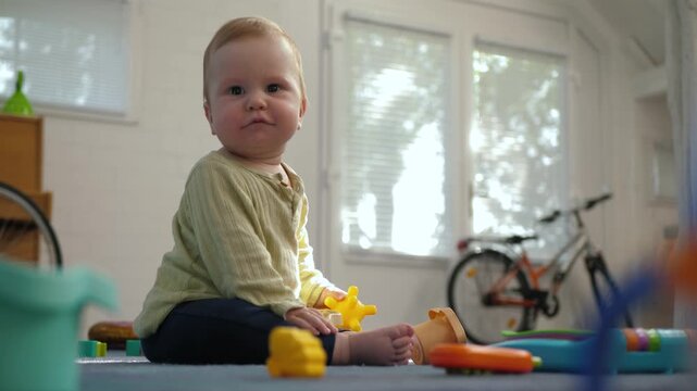 Happy baby seated on a textured playmat near the center-left of a sunlit living room, gazing upward amid scattered bright plastic toys, demonstrating curiosity and great joy