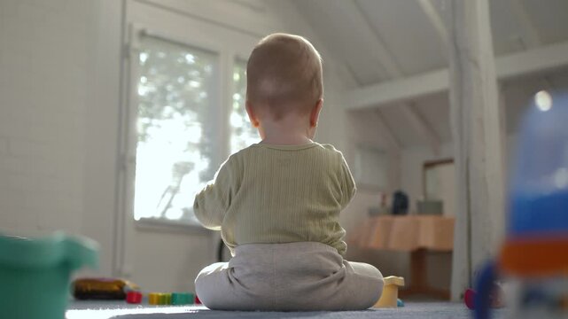 Low rear-angle shot of an infant seated on a colorful playmat in a sunlit nursery, facing a large window. Soft daylight illuminates wooden floor, emphasizing the pure innocence, childhood exploration