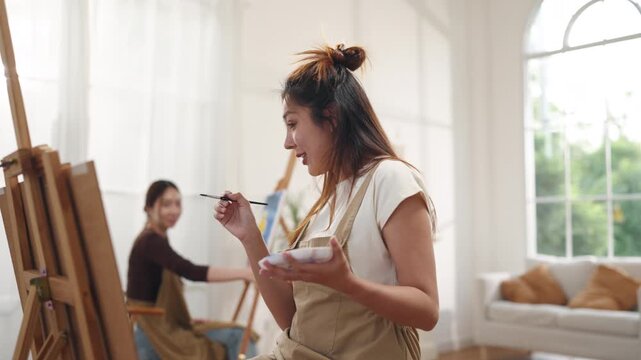 Creative young Asian women friends painting on canvas together in bright home studio. Relaxed female artists enjoying peaceful indoor leisure time, expressing imagination and passion for art.