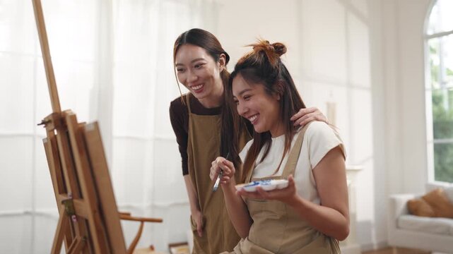 Creative young Asian women friends painting on canvas together in bright home studio. Relaxed female artists enjoying peaceful indoor leisure time, expressing imagination and passion for art.