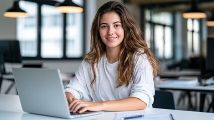 Fototapeta premium A young woman with long, wavy hair, wearing a white shirt, sitting at a desk with a laptop, smiling and looking at the camera.