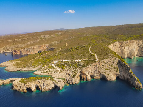 Aerial view of the rugged coastline with dramatic limestone cliffs, sea caves, turquoise waters, and a winding dirt path on the island in Zakynthos, Greece.