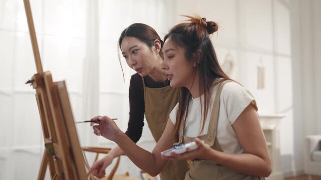 Creative young Asian women friends painting on canvas together in bright home studio. Relaxed female artists enjoying peaceful indoor leisure time, expressing imagination and passion for art.