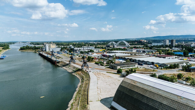Aerial view of Luka Novi Sad port facilities along the Danube River with industrial cranes, warehouses, and the Zezelj Bridge in the background in Novi Sad, Vojvodina, Serbia.