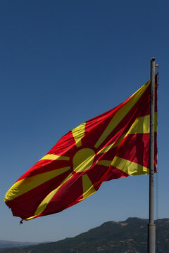 View of the national flag of North Macedonia waving on a flagpole against a clear blue sky with mountains in the background in Komuna e Ohrit, North Macedonia.