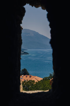 View of Lake Ohrid framed by a stone window opening, with a terracotta roof and lush trees in the foreground and mountains in the distance in Komuna e Ohrit, North Macedonia.