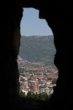 View of the town of Ohrid framed by a stone opening, with red-tiled roofs, buildings, and mountains in the background in Komuna e Ohrit, North Macedonia.
