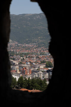 View of the town of Ohrid framed by a stone opening, with terracotta rooftops, dense trees, and mountains in the background, Komuna e Ohrit, North Macedonia.