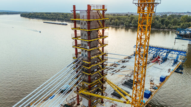 Aerial view of a bridge under construction with a tall concrete pylon, yellow crane, and steel cables spanning the Danube River in Novi Sad, Serbia.