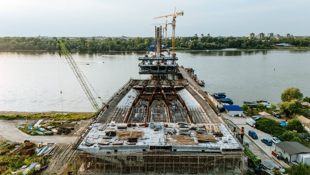 Aerial view of a bridge under construction over the Danube River with cranes and building materials on the riverbank in Novi Sad, Serbia.