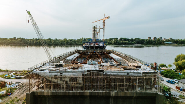Aerial view of a bridge under construction over the Danube River with cranes, steel reinforcement, and concrete structures in Novi Sad, Serbia.
