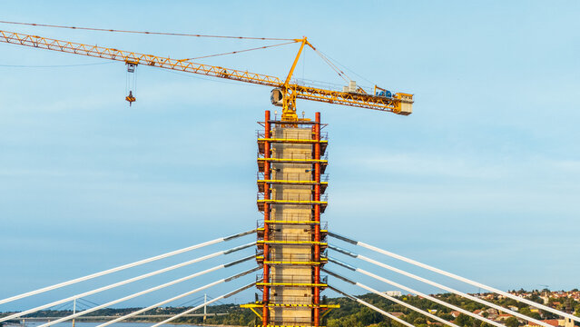 Aerial view of a bridge construction site featuring a yellow tower crane atop a concrete pylon with cable-stayed elements extending over the river in Novi Sad, Serbia.