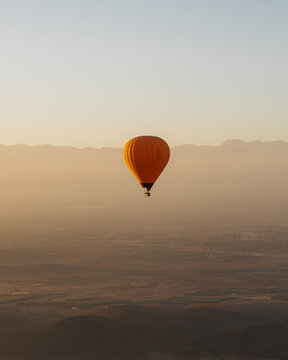 View of a vibrant orange hot air balloon floating over the vast, hazy desert landscape at sunrise near Marrakesh, Marrakesh-Safi, Morocco.