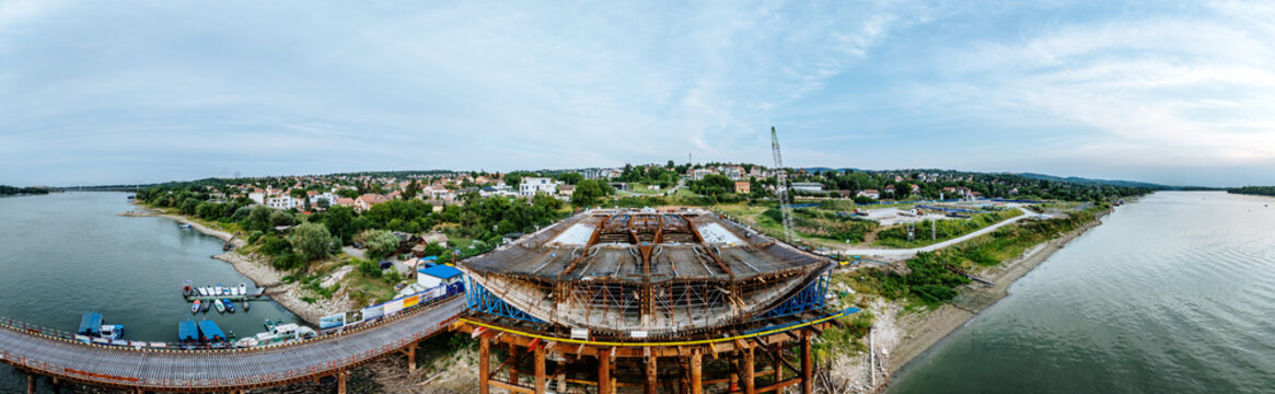 Aerial view of a bridge construction site over the Danube River with steel scaffolding, support structures, and small boats moored along the riverbank in Novi Sad, Vojvodina, Serbia.