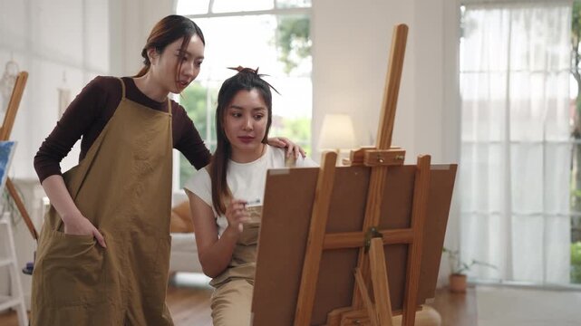 Creative young Asian women friends painting on canvas together in bright home studio. Relaxed female artists enjoying peaceful indoor leisure time, expressing imagination and passion for art.