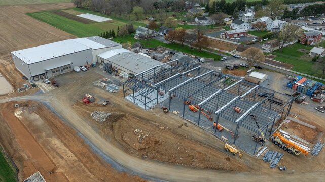 Aerial view of a construction site featuring a large steel frame structure, heavy machinery, and an adjacent warehouse building in a rural setting in USA.