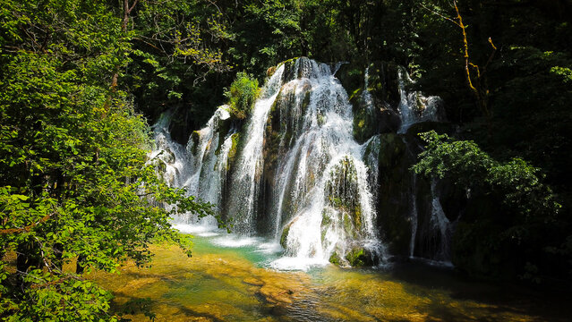 Aerial view of Cascade des Tufs waterfall cascading over mossy rocks into a clear turquoise pool surrounded by lush green forest in Les Planches-pres-Arbois, Bourgogne-Franche-Comte, France.