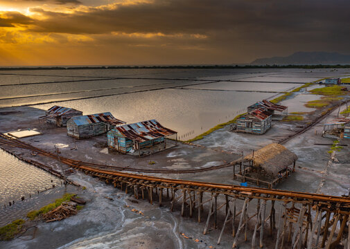 Aerial view of Salinas de Bani salt pans with rustic wooden huts, narrow rail tracks, and reflective water pools under a dramatic golden sunset sky in Bani, Peravia, Dominican Republic.