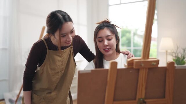 Creative young Asian women friends painting on canvas together in bright home studio. Relaxed female artists enjoying peaceful indoor leisure time, expressing imagination and passion for art.