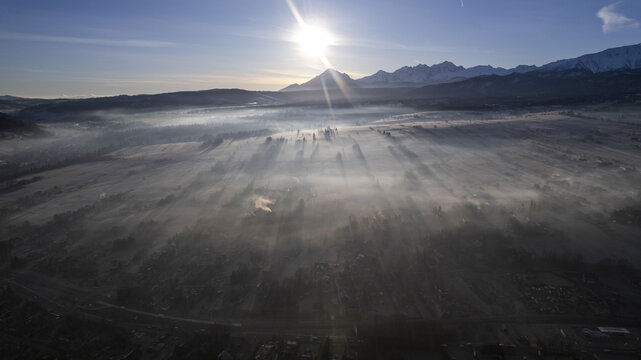 Aerial view of a misty valley at sunset with sun rays illuminating the forest and snow-capped mountains in the background in Poland.