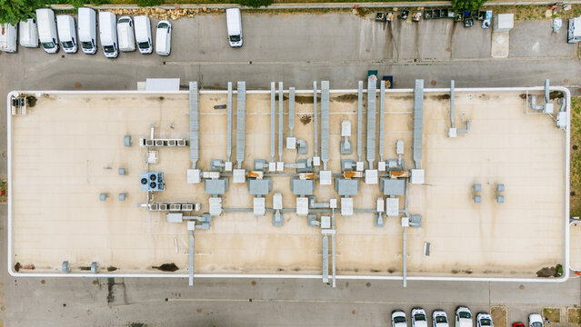 Aerial view of a flat commercial building roof with numerous HVAC units, ventilation ducts, and metal piping, with parking lots and white vans in Novi Sad, Vojvodina, Serbia.