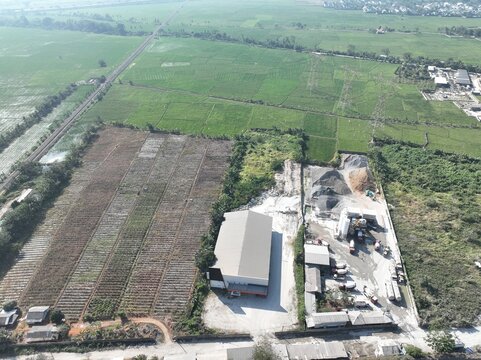 Aerial view of an industrial warehouse facility with concrete trucks and storage piles, situated next to green agricultural fields and a railway line in Cilegon, Banten, Indonesia.