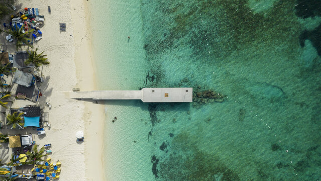 Aerial view of Junkanoo Beach with a concrete pier extending into the turquoise Caribbean Sea, featuring white sand, palm trees, and beach facilities in Nassau, New Providence, The Bahamas.