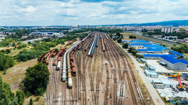 Aerial view of a busy railway yard with numerous freight trains, cargo containers, and industrial buildings under a cloudy blue sky in Novi Sad, Vojvodina, Serbia.