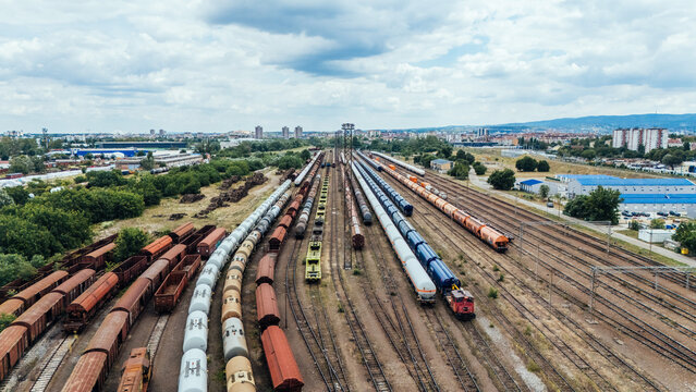 Aerial view of a large railway yard featuring numerous freight trains, tanker cars, and cargo containers on parallel tracks, with urban buildings and greenery in Novi Sad, Vojvodina, Serbia.