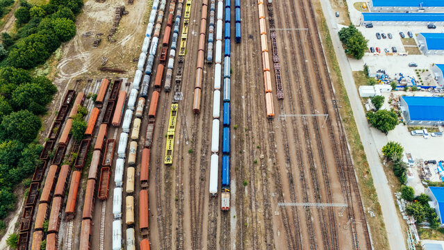 Aerial view of a busy railway yard with numerous freight train cars lined up on parallel tracks, surrounded by industrial buildings and green trees in Novi Sad, Vojvodina, Serbia.