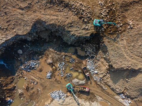 Aerial view of excavators and dump trucks working at a quarry site with piles of rocks and mud in Karanglewas, Central Java, Indonesia.