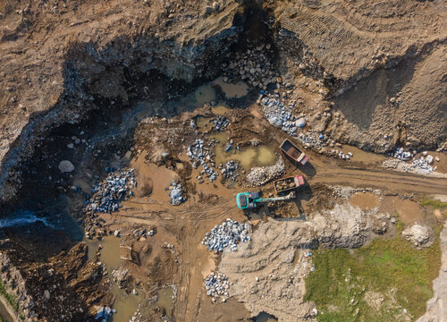 Aerial view of an active quarry site with an excavator and dump trucks working among piles of rocks, dirt, and muddy pools in Karanglewas, Central Java, Indonesia.