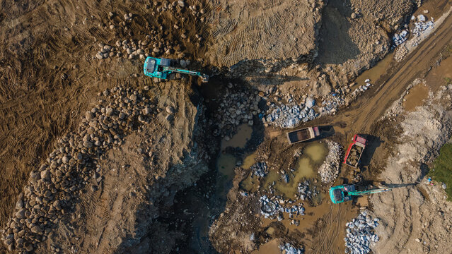 Aerial view of excavators and dump trucks working at a stone quarry with piles of rocks and muddy terrain in Karanglewas, Central Java, Indonesia.