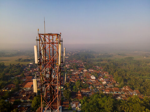 Aerial view of a telecommunications tower with antennas rising above a rural village with tiled rooftops and lush trees in Kedungbanteng, Central Java, Indonesia.
