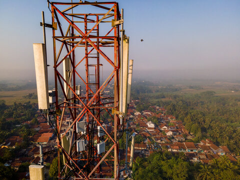 Aerial view of a telecommunications tower with antennas and metal framework overlooking a rural village with houses and lush trees in Kedungbanteng, Central Java, Indonesia.