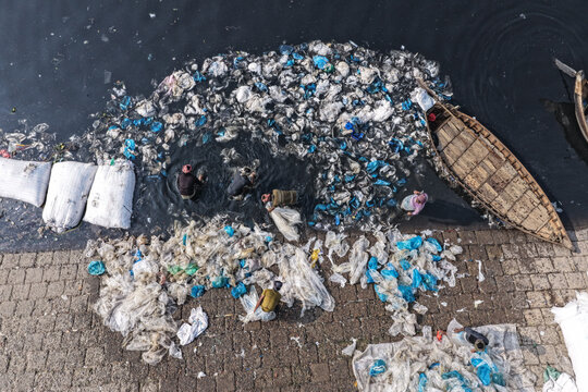 Dhaka, Bangladesh - 23 January 2026: Aerial view of workers collecting plastic waste from the Buriganga River near a stone embankment, with a wooden boat and large white sacks.