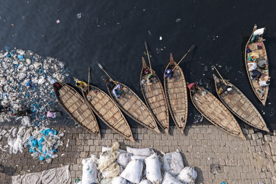 Dhaka, Dhaka Division, Bangladesh - 23 January 2026: Aerial view of wooden boats on the Buriganga River, with people on board and plastic waste floating near the stone embankment.
