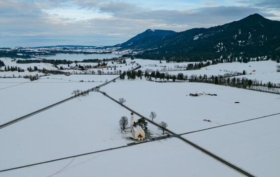 Aerial view of St. Coloman Church surrounded by snow-covered fields and trees in winter in Oberstdorf, Bayern, Germany.