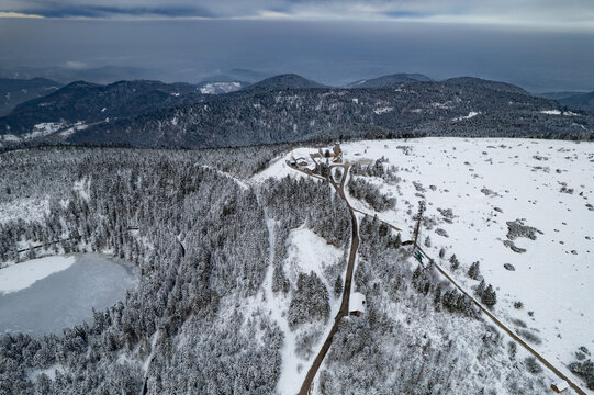 Aerial view of the snowy landscape featuring a frozen lake, dense pine forests, and a winding road leading to mountain buildings in Oberstdorf, Bayern, Germany.