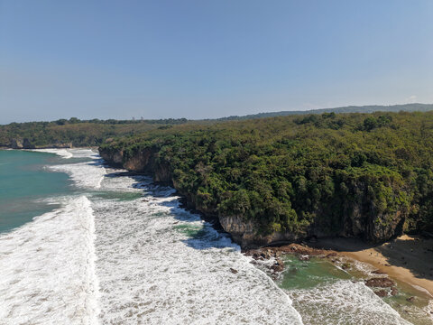 Aerial view of Pantai Goa Langir featuring rugged limestone cliffs, lush tropical forest, and white waves crashing against the shoreline in Kecamatan Bayah, Banten, Indonesia.