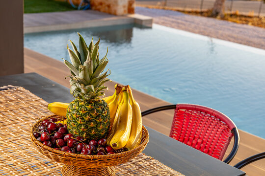 View of a basket filled with a fresh pineapple, ripe bananas, and dark red cherries on a table beside a swimming pool in Chania, Greece.