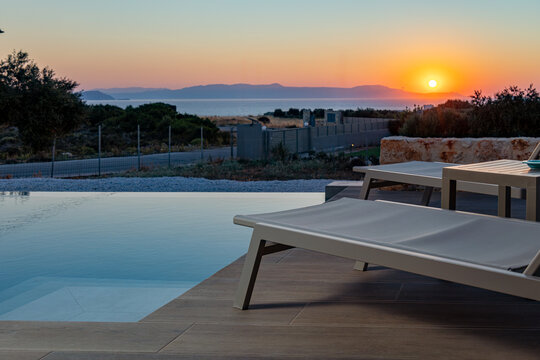 View of a modern infinity pool with lounge chairs on a wooden deck overlooking the sea during a golden sunset in Chania, Greece.