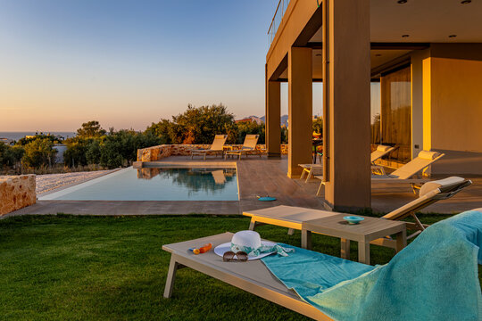 View of a modern villa terrace with an infinity pool, sun loungers, and olive trees overlooking the sea at sunset in Chania, Greece.