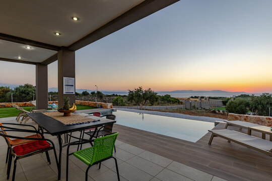 View of an infinity pool on a terrace with colorful chairs and a dining table overlooking the sea at sunset in Chania, Greece.