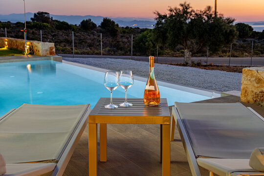 View of a bottle of rose wine and two glasses on a small table by a swimming pool at sunset with a view of the landscape in Chania, Greece.
